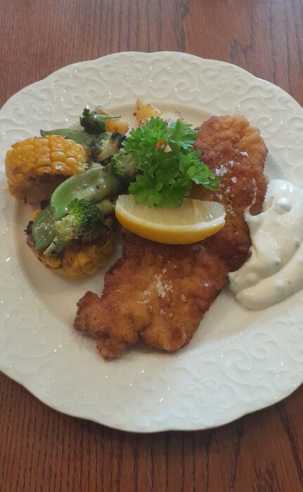 En tallrik med panerad schnitzel, grillad majs, grönsaker, citronskiva, persilja och vitlöksås på träbord.