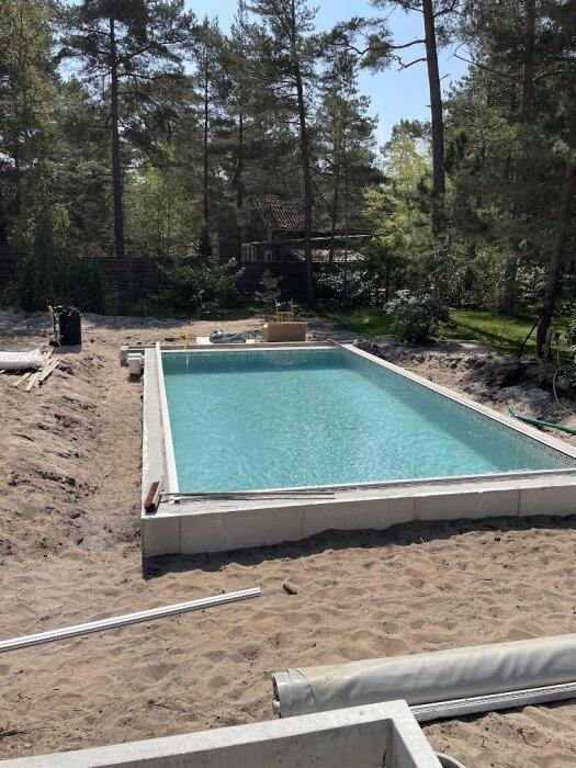 Solljus, simbassäng under konstruktion, trädgård, skogsmiljö, byggmaterial runtomkring, ofärdig pool.