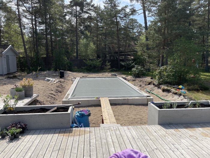 Trädgård med pool och poolskydd, trädäck, leksaker, vegetation, skjul, under konstruktion eller renovering. Solig dag.