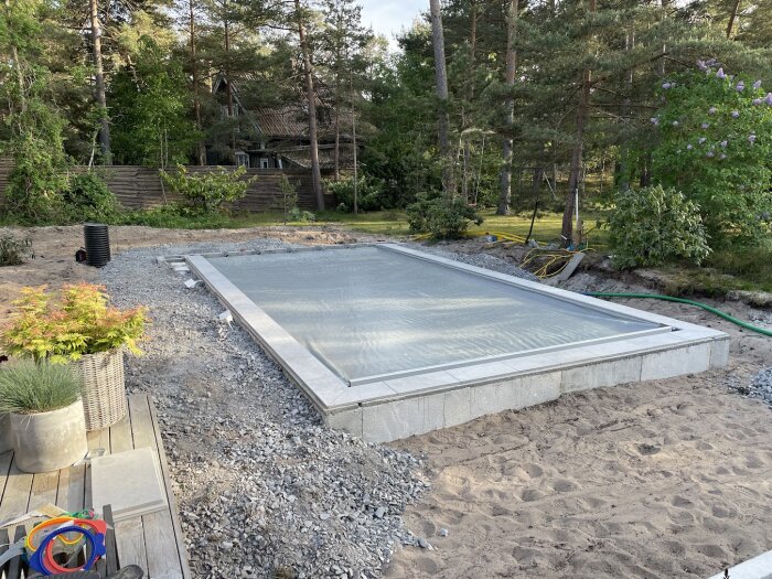 Nybyggd pool med täckning, omgiven av sand och växter, i en skogsmiljö.