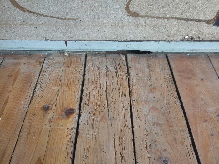 Skadat trägolv och vitmålad sockel med en spricka intill en fragmenterad vägg. Anmärkningsvärda slitage och skador synliga.