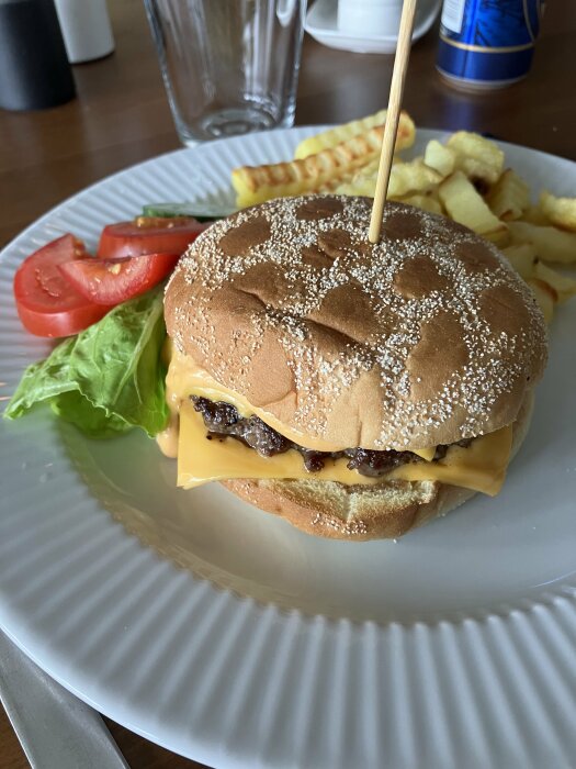 Cheeseburgare på tallrik med pommes frites, tomat, sallad, dricka i bakgrunden.