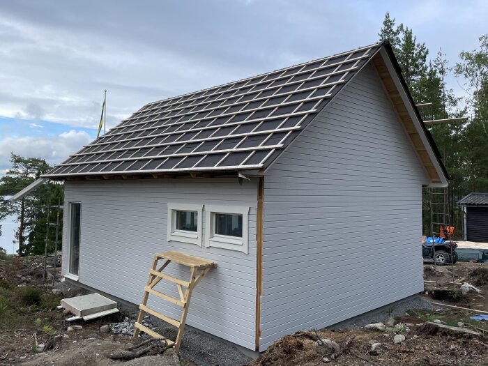 Ett nybyggt hus med tak under konstruktion, omgiven av skog och stenig mark.