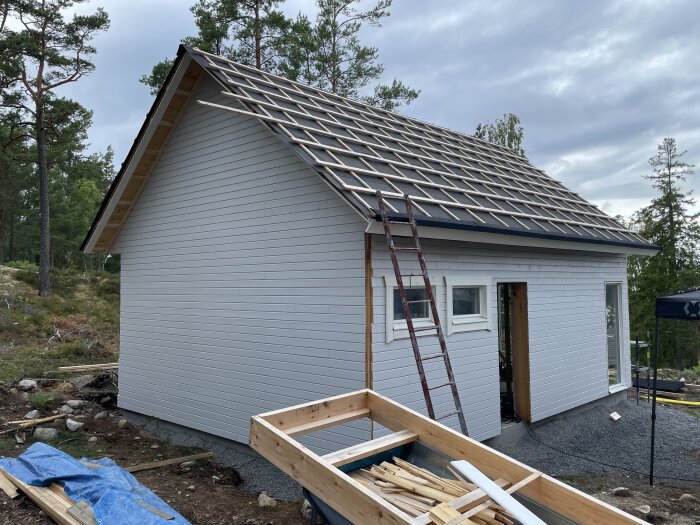 Hus under konstruktion med stege, omgivet av skog; takarbete pågår, byggmaterial synligt, molnig himmel.