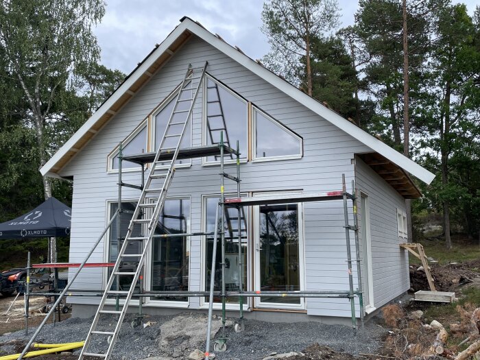 Nybyggt hus med ställningar, skogsbakgrund, grå fasad, stegar, oavslutat byggarbete.