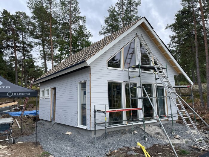 Ett vitmålat hus under konstruktion med träpanel, stegar, ställning och grus i förgrunden. Skog i bakgrunden.