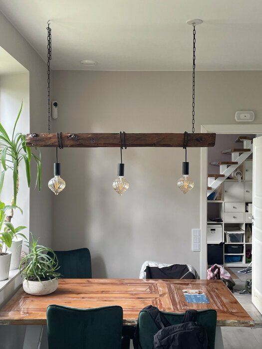 Ett träbord, gröna stolar, taklampa med träbjälke, inomhusväxt, ryggsäckar, minimalistisk stil, vitt rum.