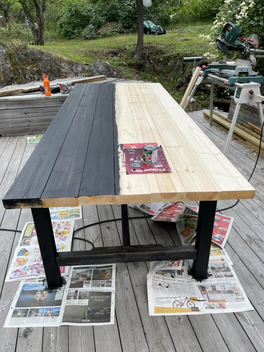 Halvmålat träbord ute, svart ben, verktyg, såg, tidningar på trädäck, grönska i bakgrund.