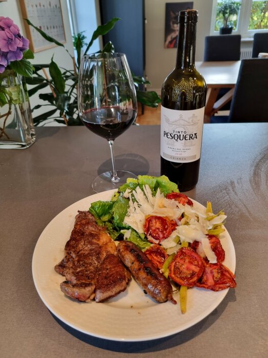 En tallrik med grillat kött, korv, sallad, rostade tomater; ett glas rött vin och en vinflaska bredvid.