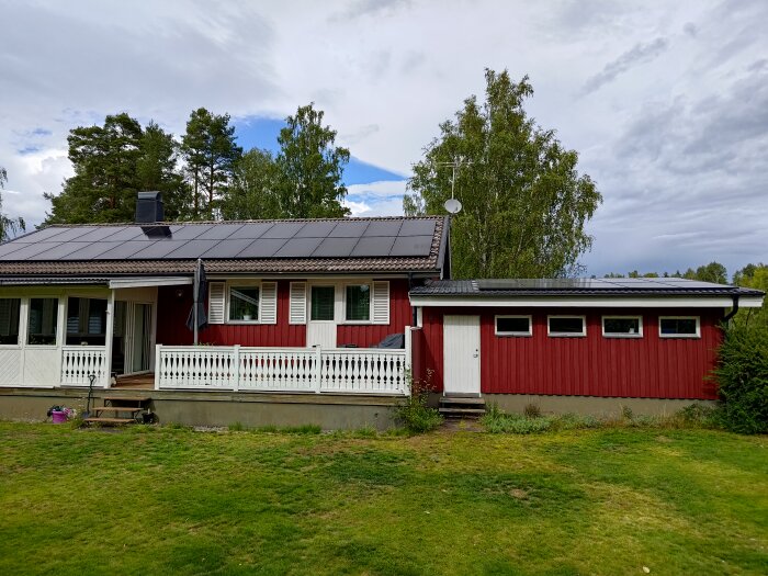 Ett rött hus med vita knutar, veranda, solpaneler på taket och grönt gräs framför.