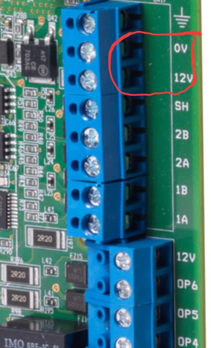 Kretskort med blå skruvplintar och etiketterade anslutningar för 0V och 12V, elektronikkomponenter synliga.