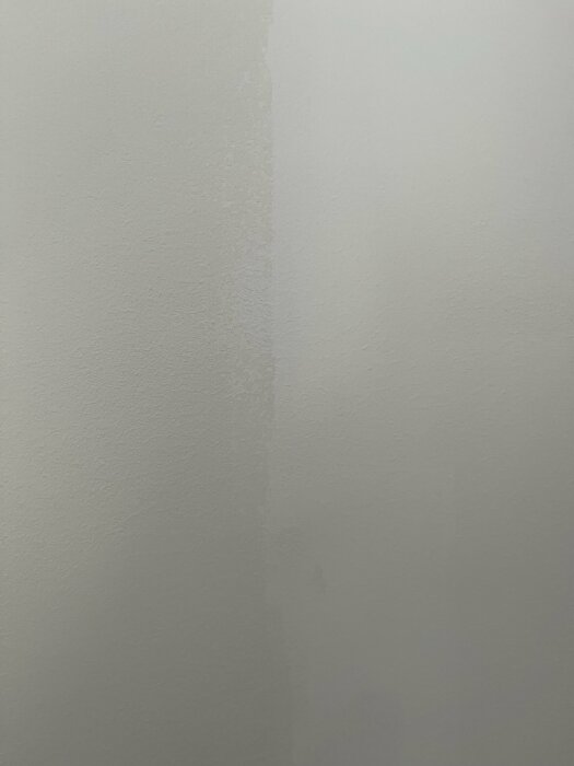 Enkel vit vägg med en synlig skarv eller möjlig vattenskada. Texturerad yta med mjuk skuggning och diffust ljus.
