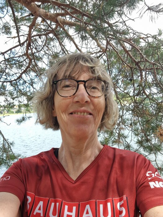 En leende person med glasögon framför träd och vatten i en röd tröja med text.