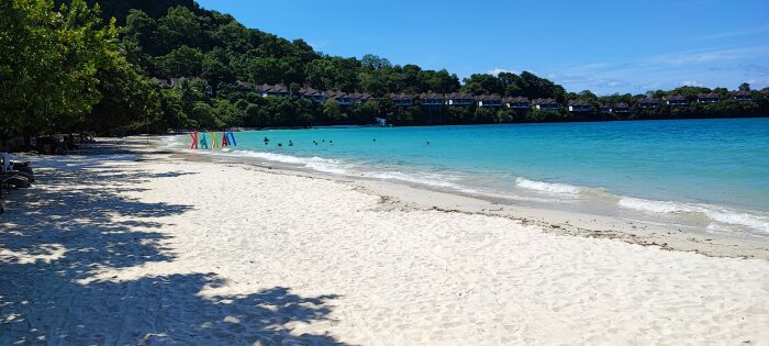 Sandy beach, clear blue water, bathers, tropical trees, sunny day, beachfront villas on hillside, calm sea.