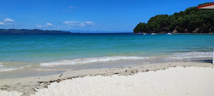 Tropisk strand, klarblått vatten, vit sand, båtar, bergig ö, soligt, blå himmel.