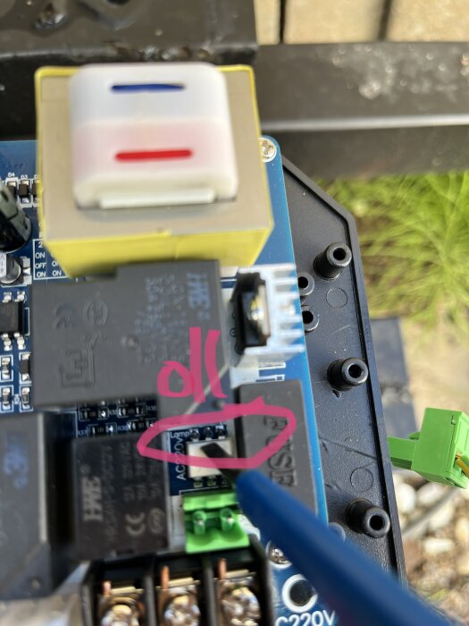 Elektronikkort med reläer, terminaler och USB-port, markerat med rosa kritsnål, suddig bakgrund.