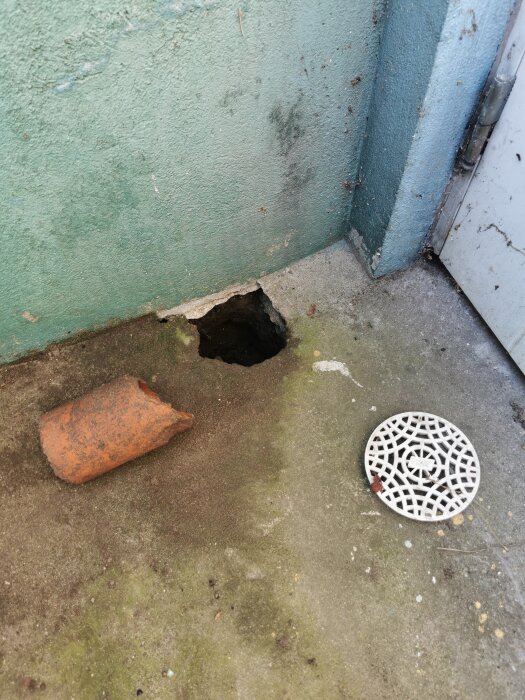 Hål i marken vid vägg, rostig tegelsten, vit avloppslock på betongyta.