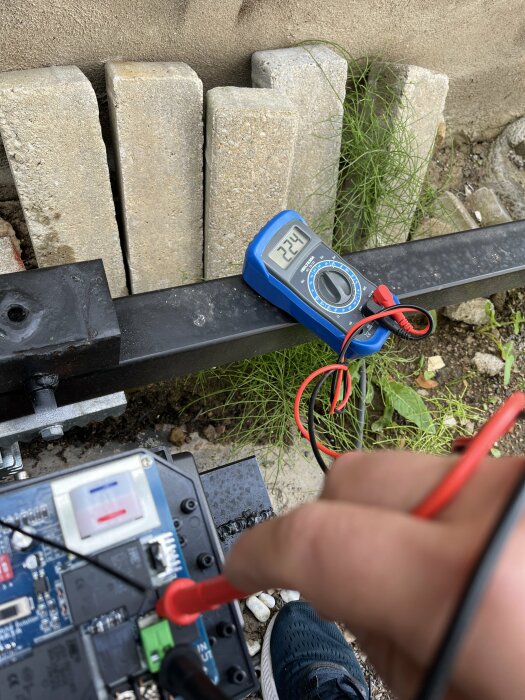 Elektroniskt projekt utomhus; multimeter visar spänning; kablar kopplade till kretskort; betongstenar och gräs i bakgrunden.