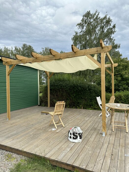 Trädäck med pergola, markis, hopfällt bord, stolar och en shoppingväska utomhus.