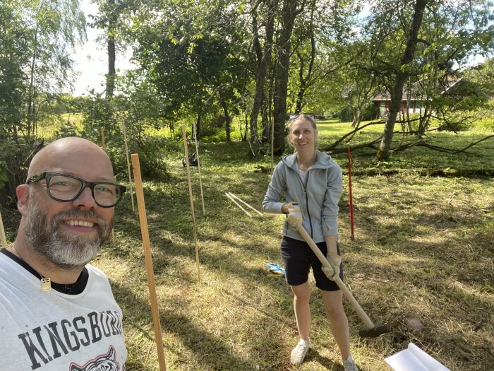 Två personer i trädgården, arbetar utomhus, soligt, grönt gräs, träd, glada ansikten, verktyg för trädgårdsarbete, röd mätpinne.