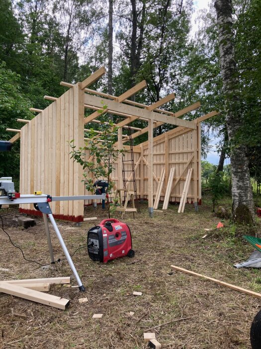 Träkonstruktion i skogsmiljö, troligen bygge av hus eller förråd, verktyg och sågbock synliga, byggprocess, naturbakgrund.