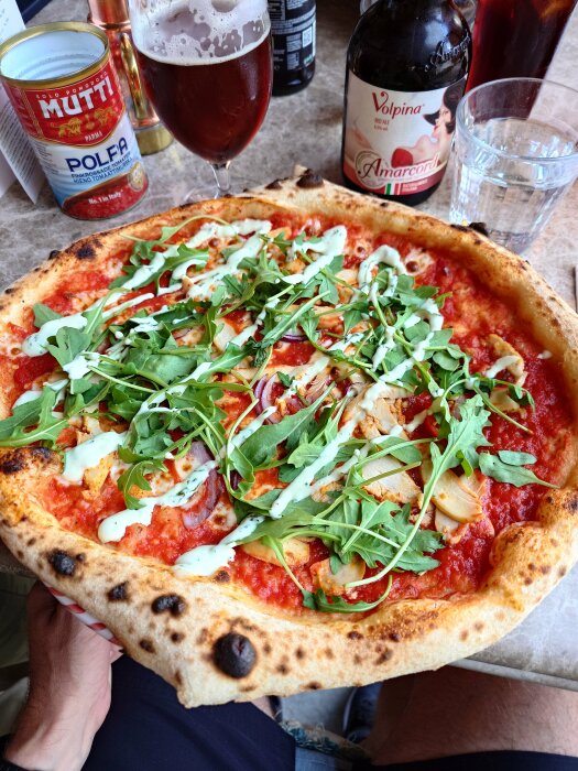 Pizza med ruccola, ost och tomatsås, öl, krossade tomater, vattenglas, sittande person.