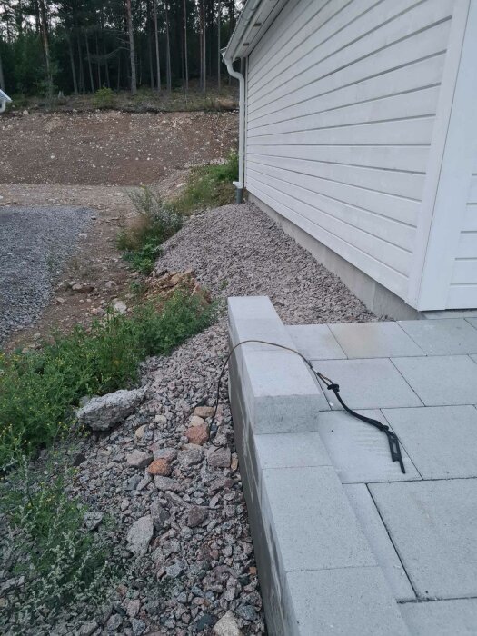 Hustavla, skogsbryn, grus och sten, grå markstenar, svart rem på vit betongvägg.