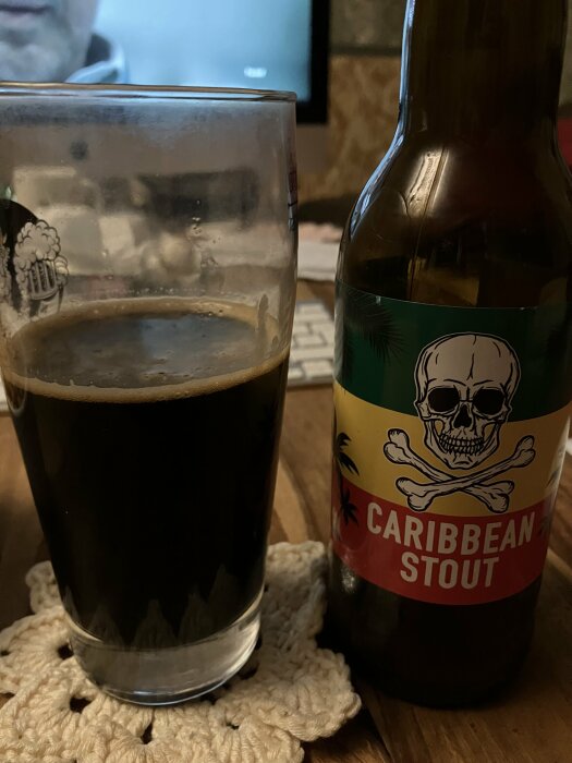 Mörkt stoutöl i glas bredvid flaska med etikett "Caribbean Stout", dödskalle, på stickad duk.