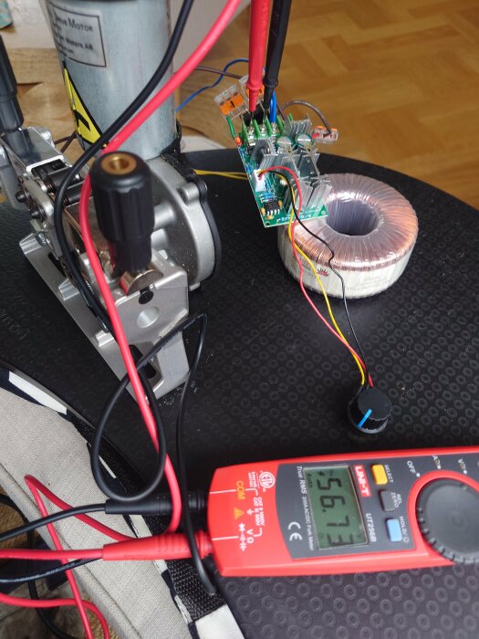 Elektrisk motor, kretskort, induktorspole, multimeter visar volt, ingenjörsprojekt eller experiment på svart underlag.