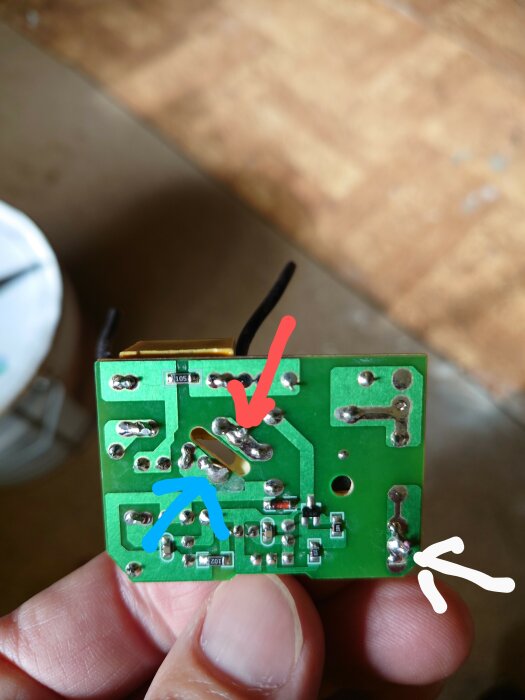 Hand håller elektroniskt kretskort med markeringar, potentiella lötproblem eller komponentpositioner, otydlig bakgrund.
