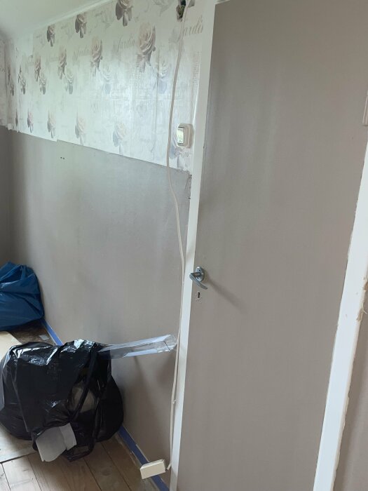 Ett hörn av ett rum under renovering med tapet, eluttag, dörr och en svart sopsäck.