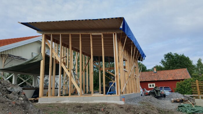 Konstruktion av trästomme till byggnad, omgivet av byggmaterial, mot bakgrund av bostadshus och grönska.