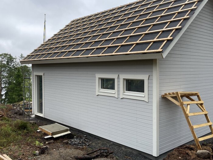 Nybyggt hus med träpanel, stegar, takläggning pågår, svenska flaggan i bakgrunden, fönster, grå himmel.