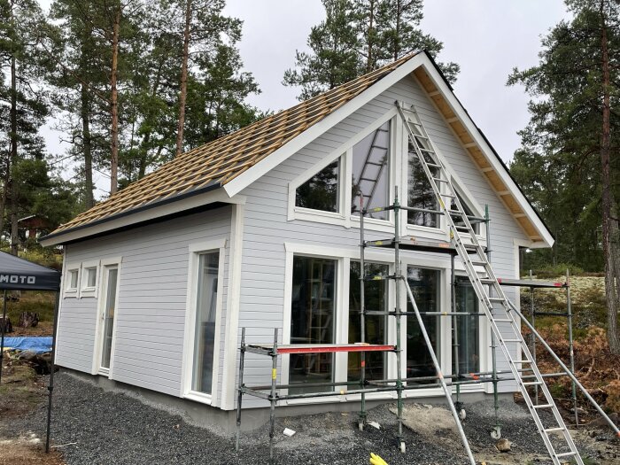 Hus under konstruktion, vita väggar, takstolar synliga, skogsbakgrund, byggställning, stege, fönster, grusgrund.