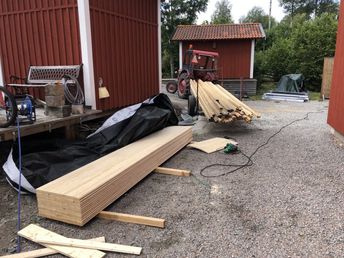 Byggplats med timmer, röd traktor, byggnadsmaterial och verktyg ute på grusgård.