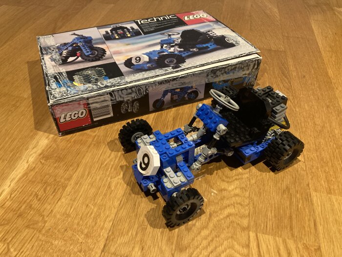 LEGO Technic modell av en blå bil framför dess öppnade förpackning på trägolv.