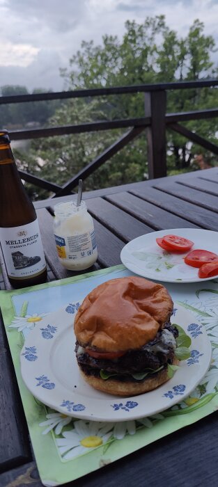 Utomhusmåltid med öl, hamburgare, majonnäs, tomat på ett träbord, omgiven av natur.