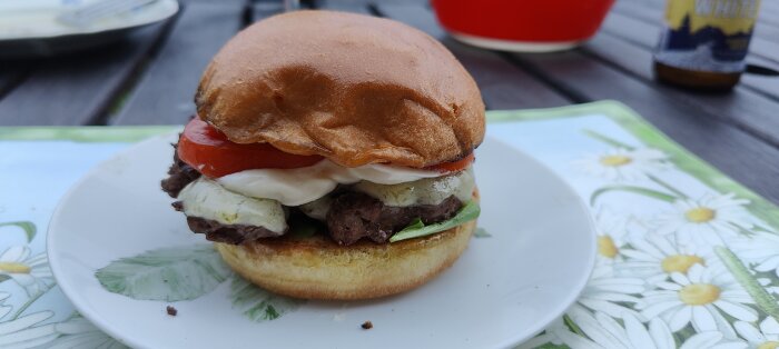 En saftig hemlagad hamburgare på tallrik med tomater, sallad och dressing, utomhus nära en dryck.