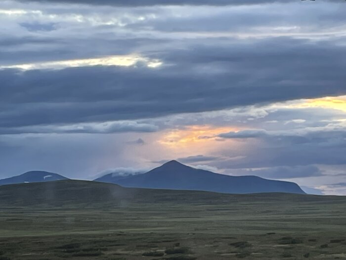 Mystisk landskap med bergssiluetter, dramatiska moln och en antydan av solnedgång i fjärran.