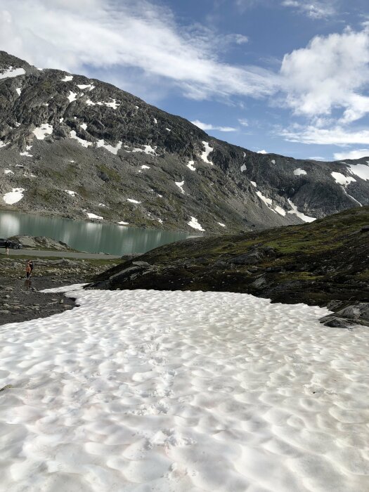 Snötäckt terräng framför berg och sjö under en blå himmel, med en person i bakgrunden.