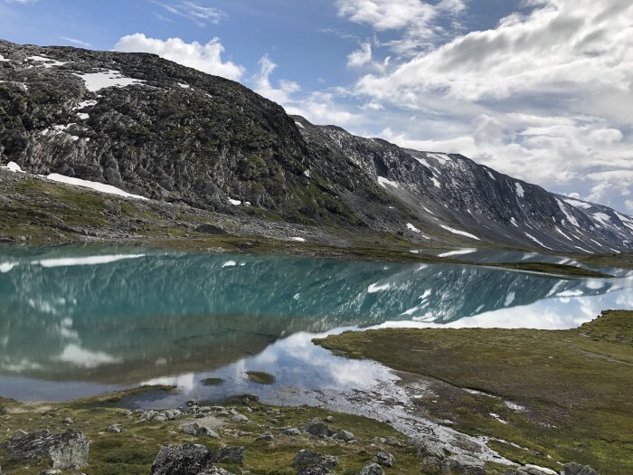 Turkost glaciärvatten speglar berg och himmel i en lugn fjällmiljö med snöfläckar och klippor.