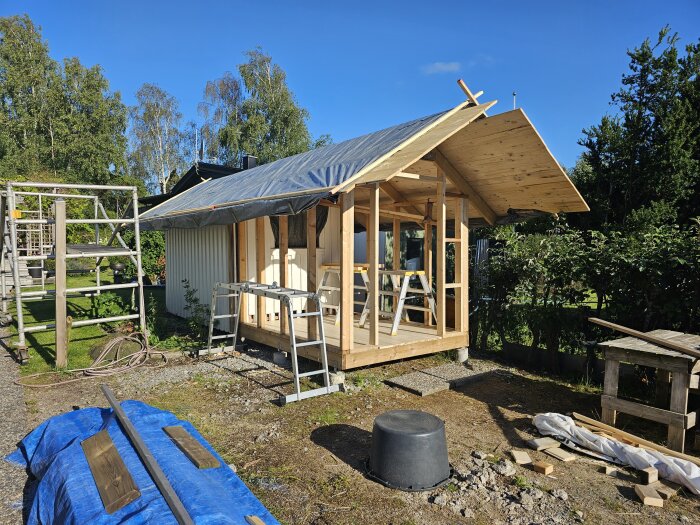 Ett halvfärdigt trähus under konstruktion med byggmaterial och verktyg utspridda på marken.