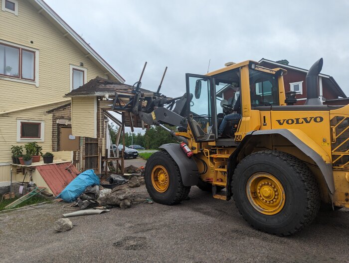 Gul Volvo hjullastare vid hus under rivning eller renovering, rörigt med bråte runt omkring.