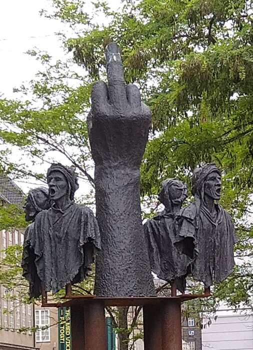 Skulptur av en handgest och fyra figurer med uttrycksfulla ansikten, konstinstallation utomhus, träd i bakgrunden.