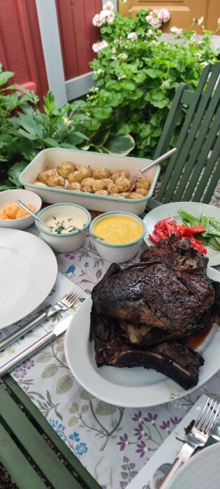 Utomhusmiddag med grillat kött, potatis, såser och grönsaker på ett blommigt dukat bord.