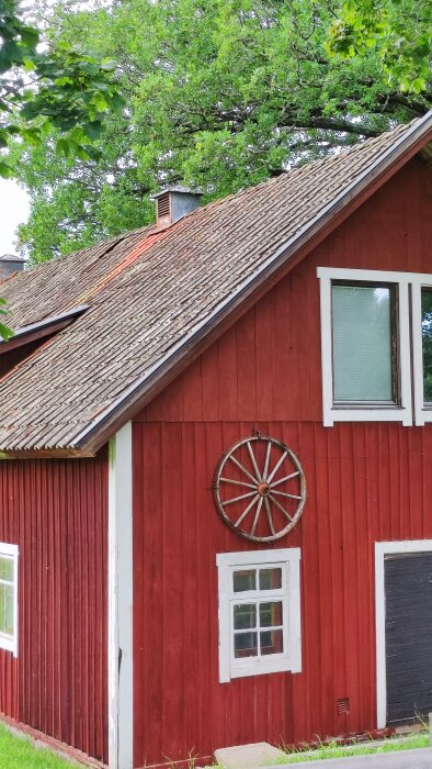 Rött trähus med tegeltak, vita fönster och ett vagnshjul på väggen. Träd i bakgrunden.