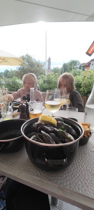 Två barn vid ett utomhusbord med musslor, bröd, kryddkvarn och dryckesglas.