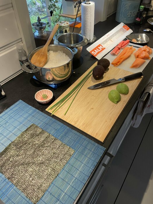 Kök med ingredienser för sushi: avokado, lax, ris, gräslök, lime, sjögrästallrik på bambumatta.