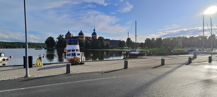 Marina med båtar framför historisk byggnad, blå himmel med moln, solglans, reflekterande vatten, gatlyktor, gröna träd.