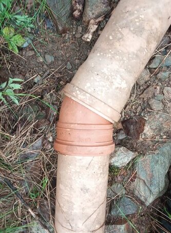 Brun PVC-koppling ansluter två delar av ett beige avloppsrör, omgivet av sten och vegetation.